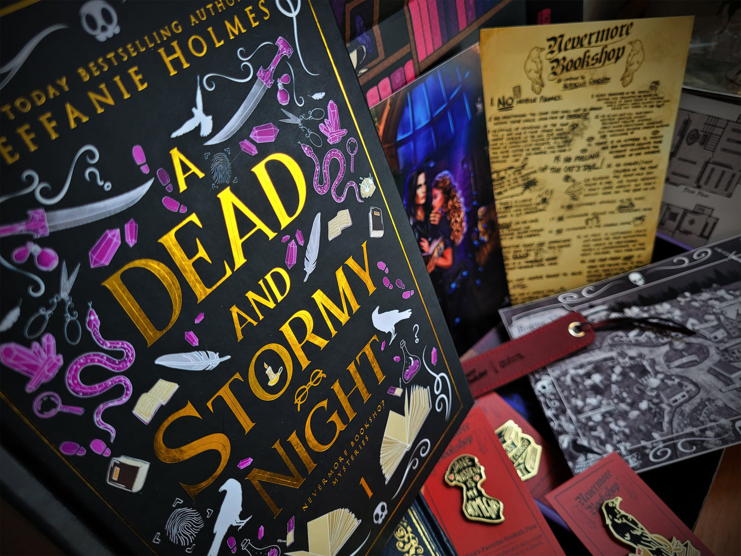 A collection of items from the Nevermore Bookshop Mysteries Book Box, including a signed hardcover of 'A Dead and Stormy Night', various art prints, enamel pins, a leatherette bookmark, and stickers, all themed around the book's content.