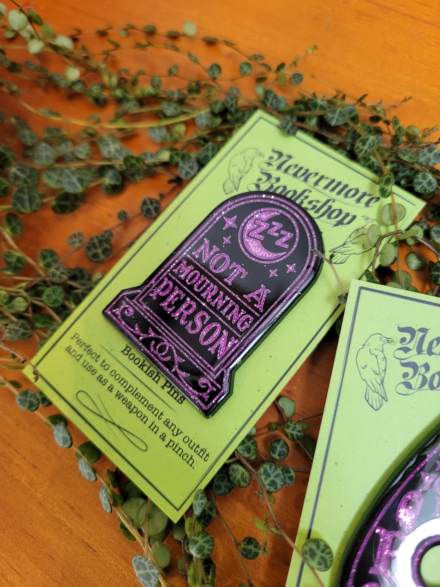 'Not a mourning person' enamel pin based on the Grimdale Graveyard Mysteries, displayed on a green card with a decorative border. The pin is black and purple in color.