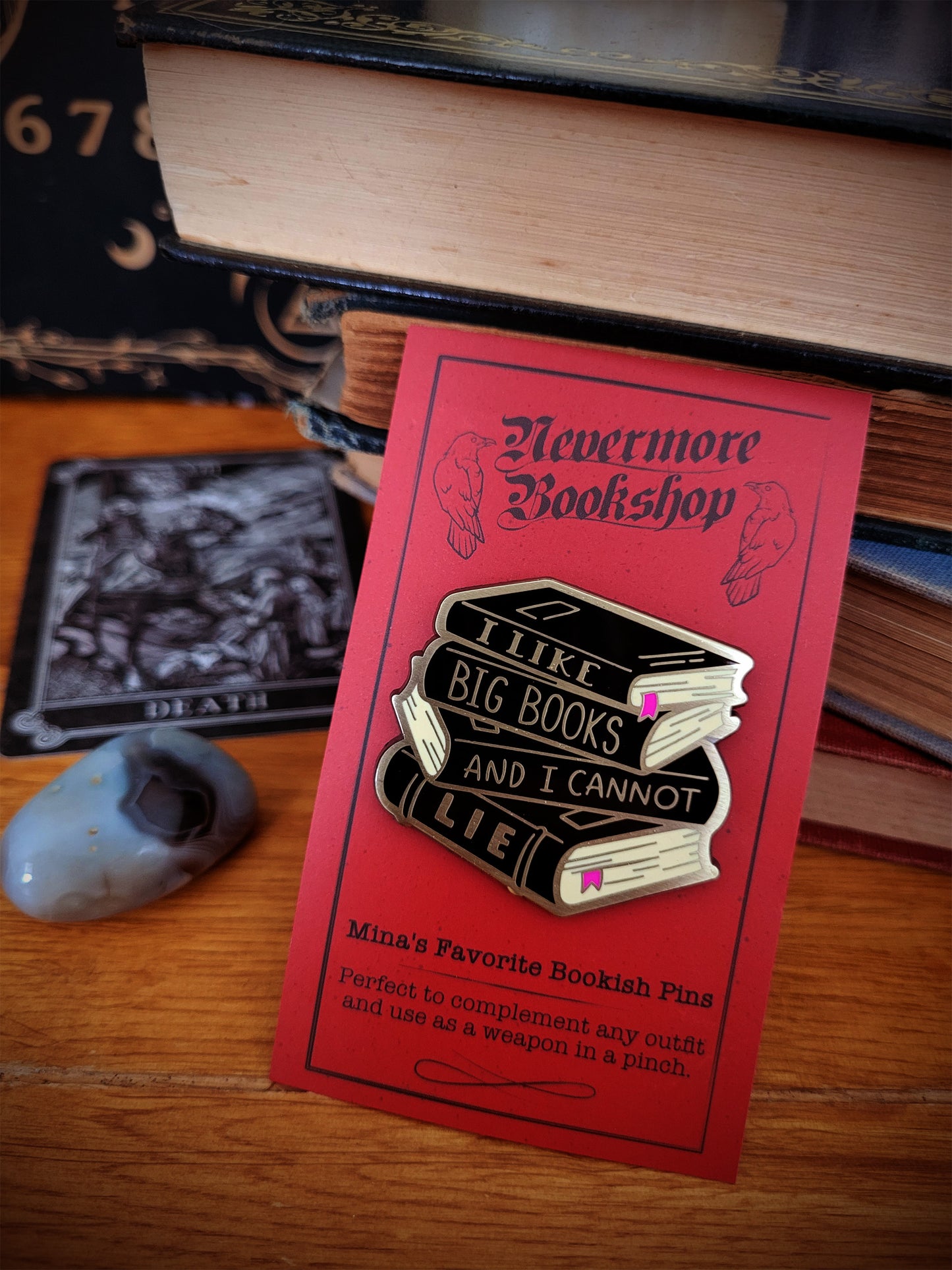 Enamel pin with designs related to the Nevermore book series, displayed on a wooden surface next to a key and a Ouija board.