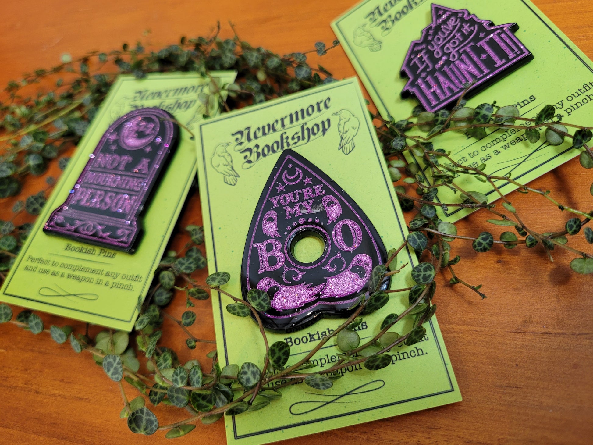 Three enamel pins with designs based on the Grimdale Graveyard Mysteries, displayed on green cards with a decorative border. The pins are black and purple in color.