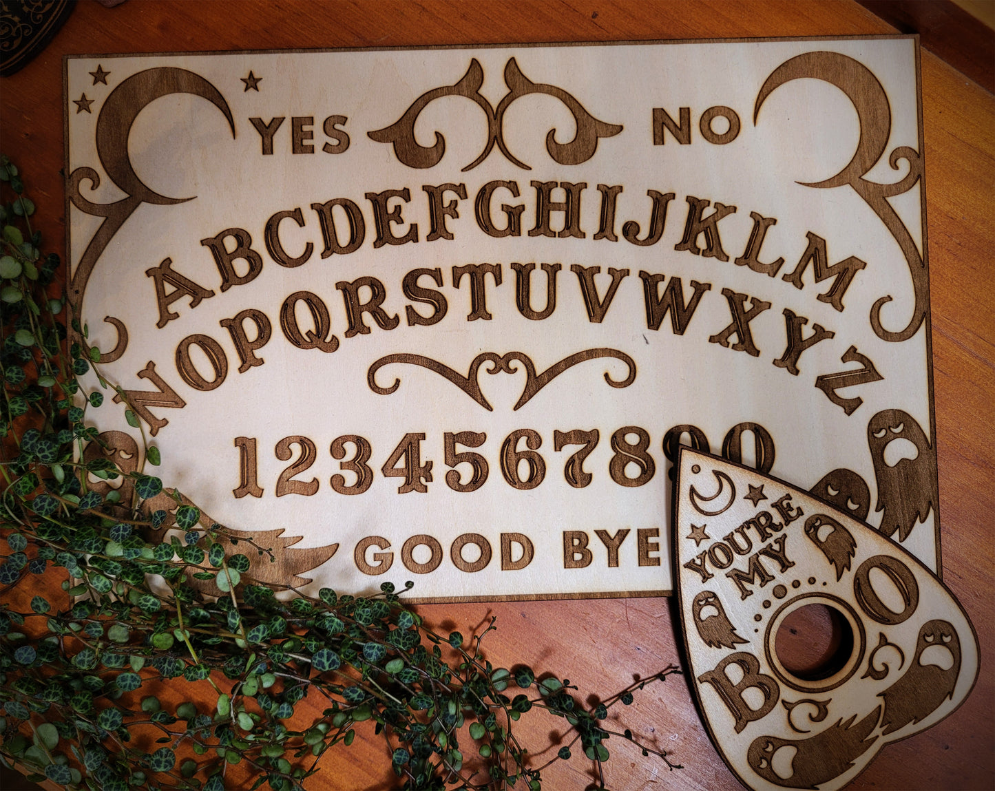 A wooden spirit board with laser-engraved letters, numbers, and designs, accompanied by a planchette with a hole in the center.