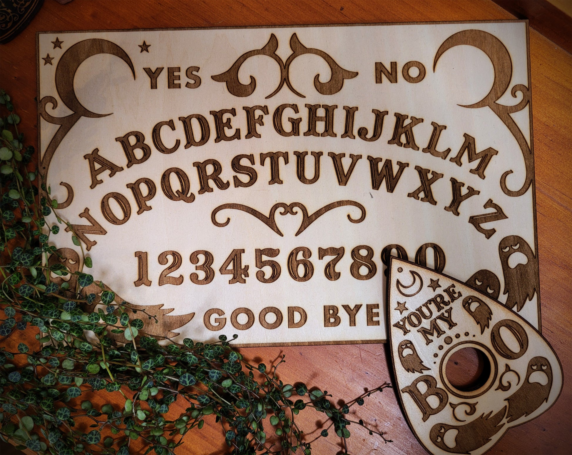 A wooden spirit board with laser-engraved letters, numbers, and designs, accompanied by a planchette with a hole in the center.