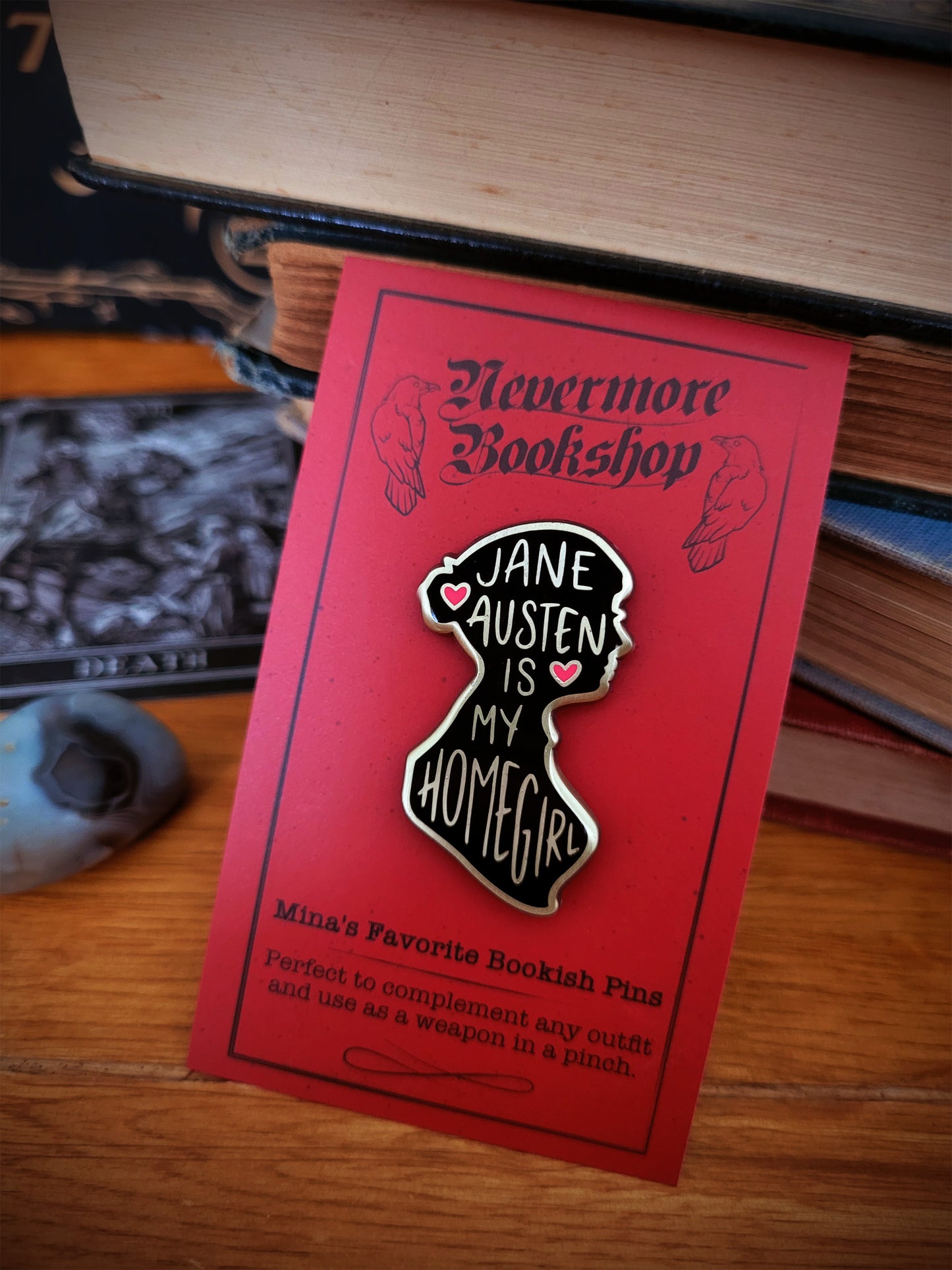 Enamel pins with designs related to the Nevermore book series, displayed on a wooden surface next to a key and a Ouija board.