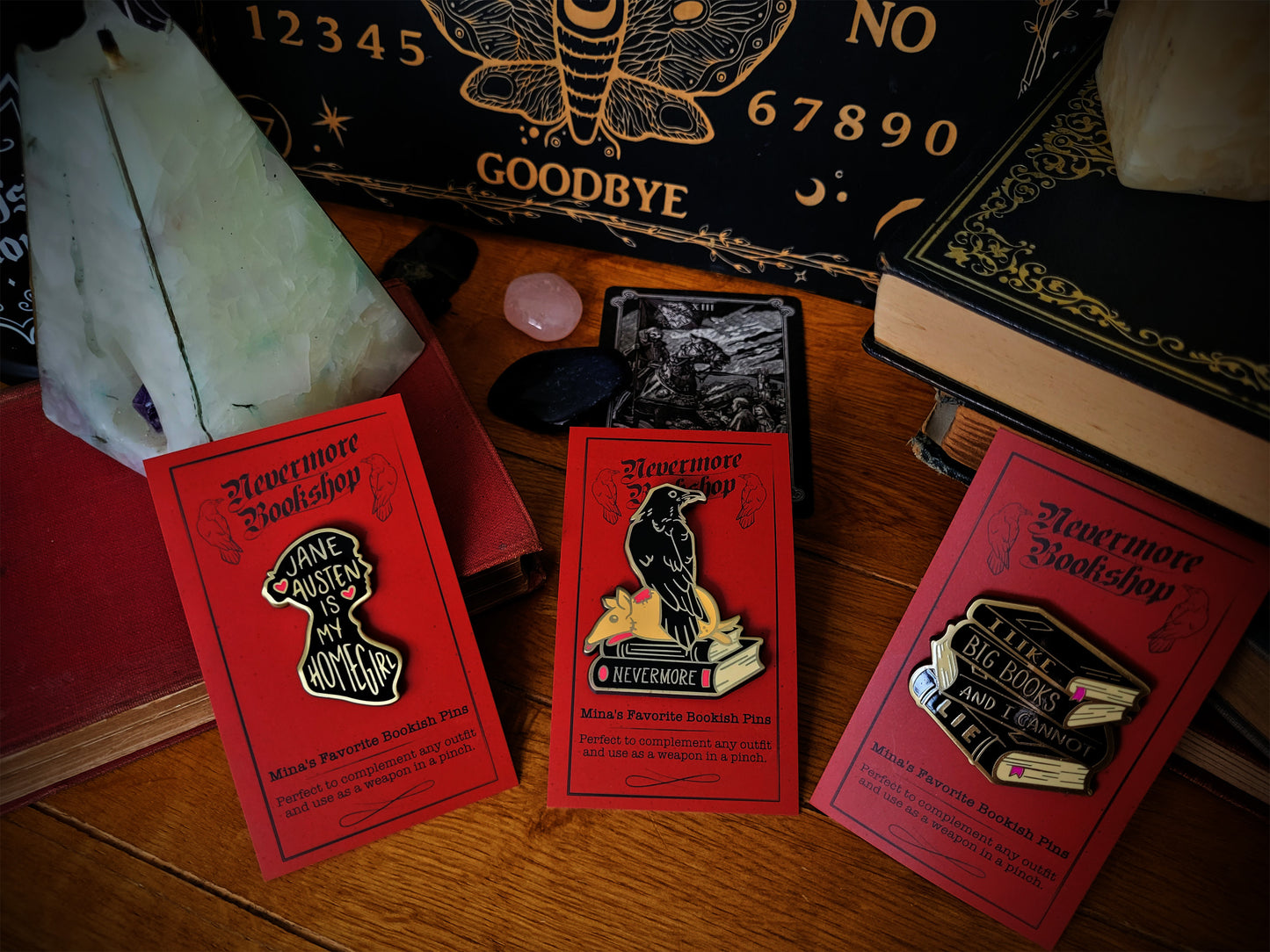 Three enamel pins with designs related to the Nevermore book series, displayed on a wooden surface next to a key and a Ouija board.