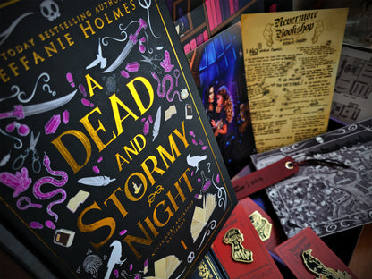 A collection of items from the Nevermore Bookshop Mysteries Book Box, including a signed hardcover of 'A Dead and Stormy Night', various art prints, enamel pins, a leatherette bookmark, and stickers, all themed around the book's content.