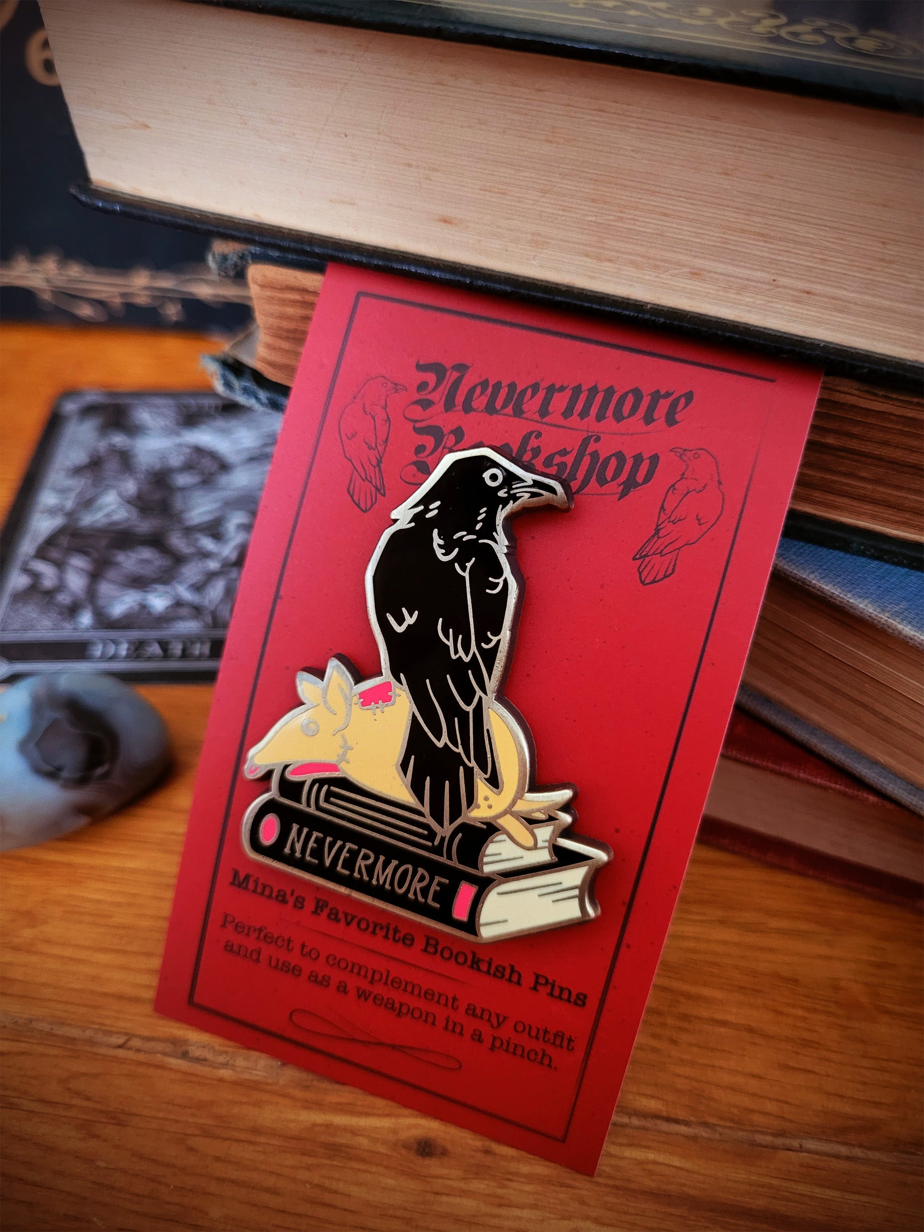 Enamel pins with designs related to the Nevermore book series, displayed on a wooden surface next to a key and a Ouija board.