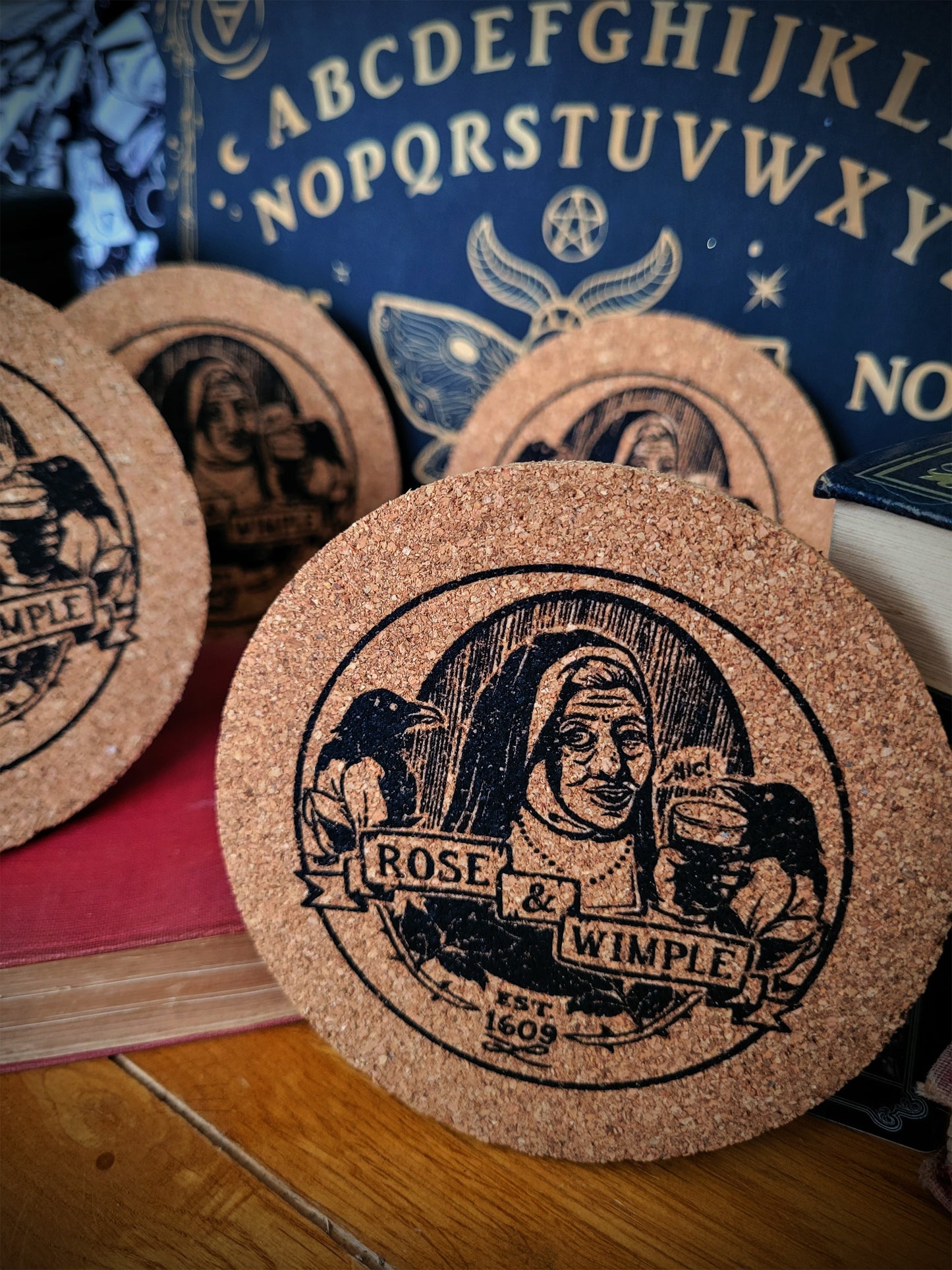 A set of round cork coasters with a black and beige Rose & Wimple design, displayed on a surface.