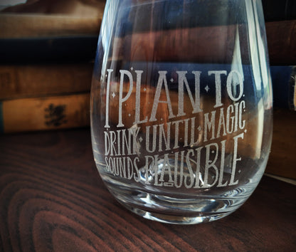 A clear wine glass with etched text that reads 'I PLAN TO DRINK UNTIL MAGIC SOUNDS PLAUSIBLE'