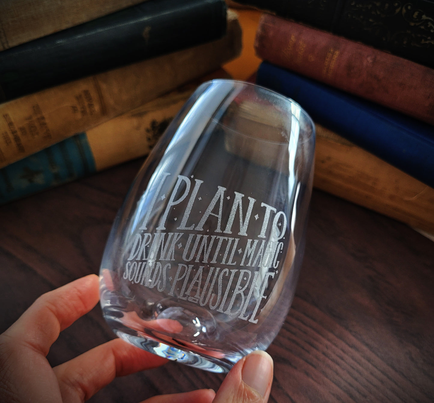 A clear wine glass with etched text that reads 'I PLAN TO DRINK UNTIL MAGIC SOUNDS PLAUSIBLE'