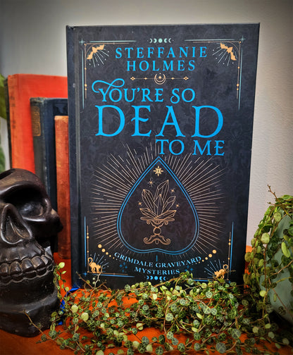 A hardcover copy of the book 'You're So Dead To Me' by Steffanie Holmes displayed on a shelf with decorative items around it. The book has a black cover with gold and white text and a decorative design.