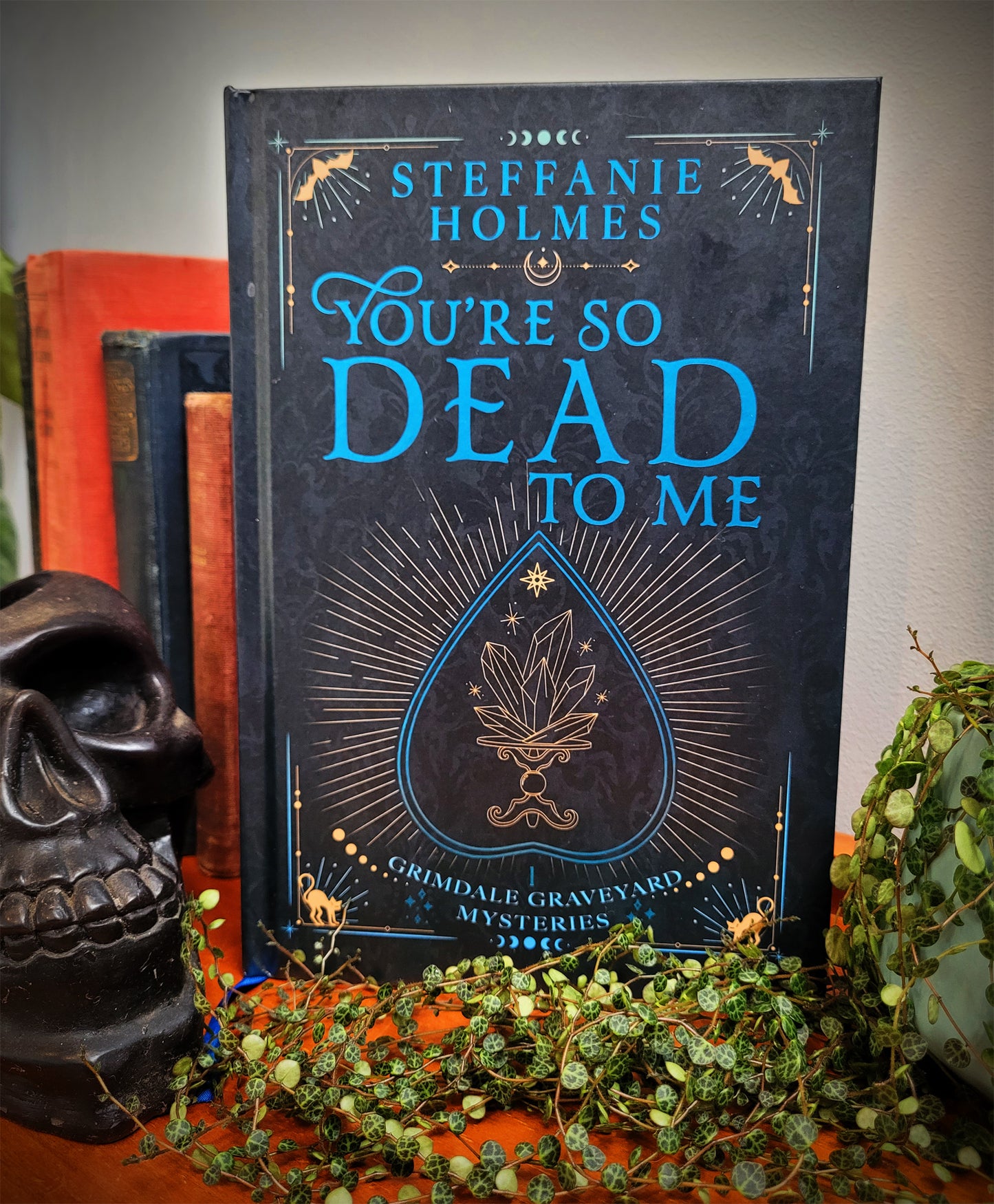 Signed hardcover edition of 'You're So Dead To Me' by Steffanie Holmes, featuring a black cover with gold and teal accents, and illustrations, Pictured with books, a skull and a plant