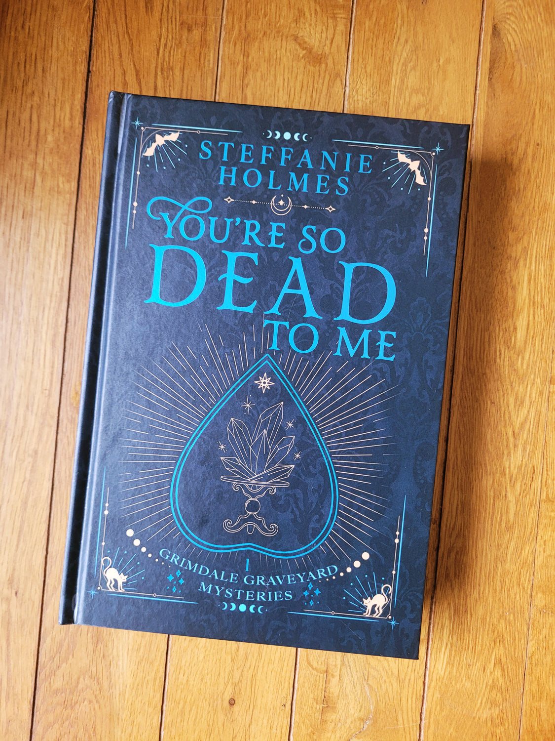 Signed hardcover edition of 'You're So Dead To Me' by Steffanie Holmes, featuring a black cover with gold and teal accents, and illustrations 