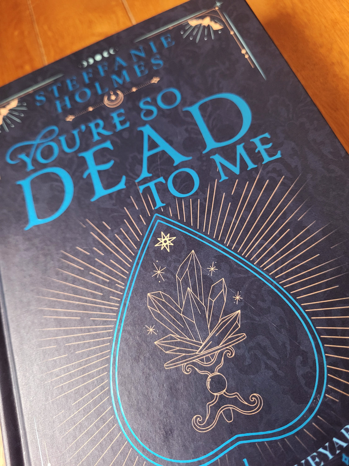 Signed hardcover edition of 'You're So Dead To Me' by Steffanie Holmes, featuring a black cover with gold and teal accents, and illustrations 